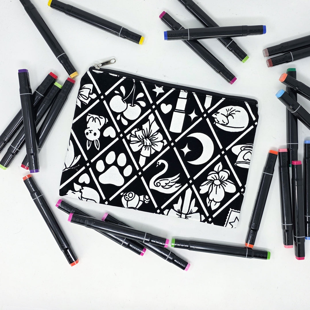 Photo of a black and white flat pencil pouch with lots of colorful markers beside it indicating the customer gets to color it when they receive theirs. This is the same bag as the pencil case and makeup bag - Side B - this bag has images of Coquette-inspired illustrations including a swan, a paw print, bows, a moon, stars, a bunny on it cherries and more