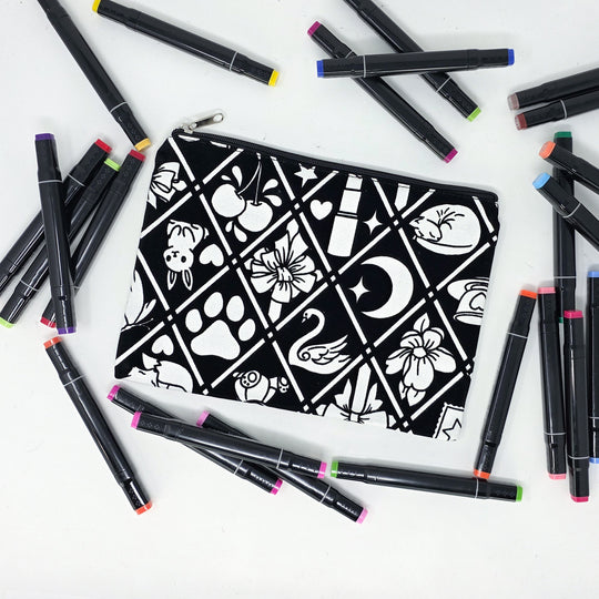 Photo of a black and white flat pencil pouch with lots of colorful markers beside it indicating the customer gets to color it when they receive theirs. This is the same bag as the pencil case and makeup bag - Side B - this bag has images of Coquette-inspired illustrations including a swan, a paw print, bows, a moon, stars, a bunny on it cherries and more
