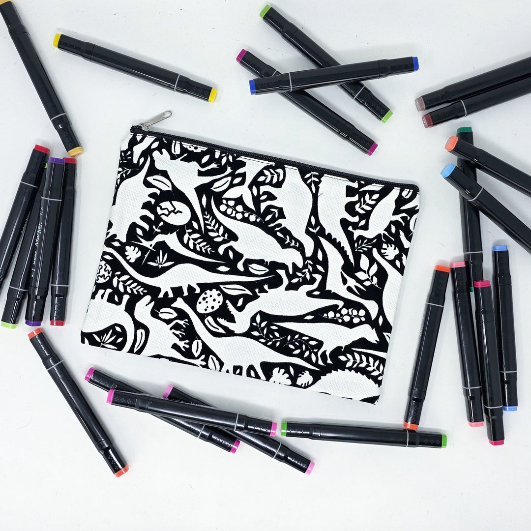 Photo of a black and white flat pencil pouch with lots of colorful markers beside it indicating the customer gets to color it when they receive theirs. This is the same bag as the pencil case and makeup bag - Side A - this bag has images of dinosaurs, foliage, and eggs