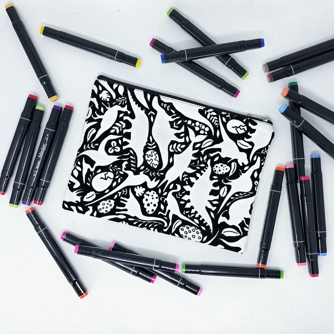Photo of a black and white flat pencil pouch with lots of colorful markers beside it indicating the customer gets to color it when they receive theirs. This is the same bag as the pencil case and makeup bag - Side A - this bag has images of dinosaurs, foliage, and eggs
