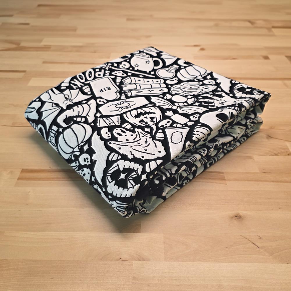 Uncolored Spooky Halloween blanket with ghosts, skulls, pumpkins, and other themed images folded on a table
