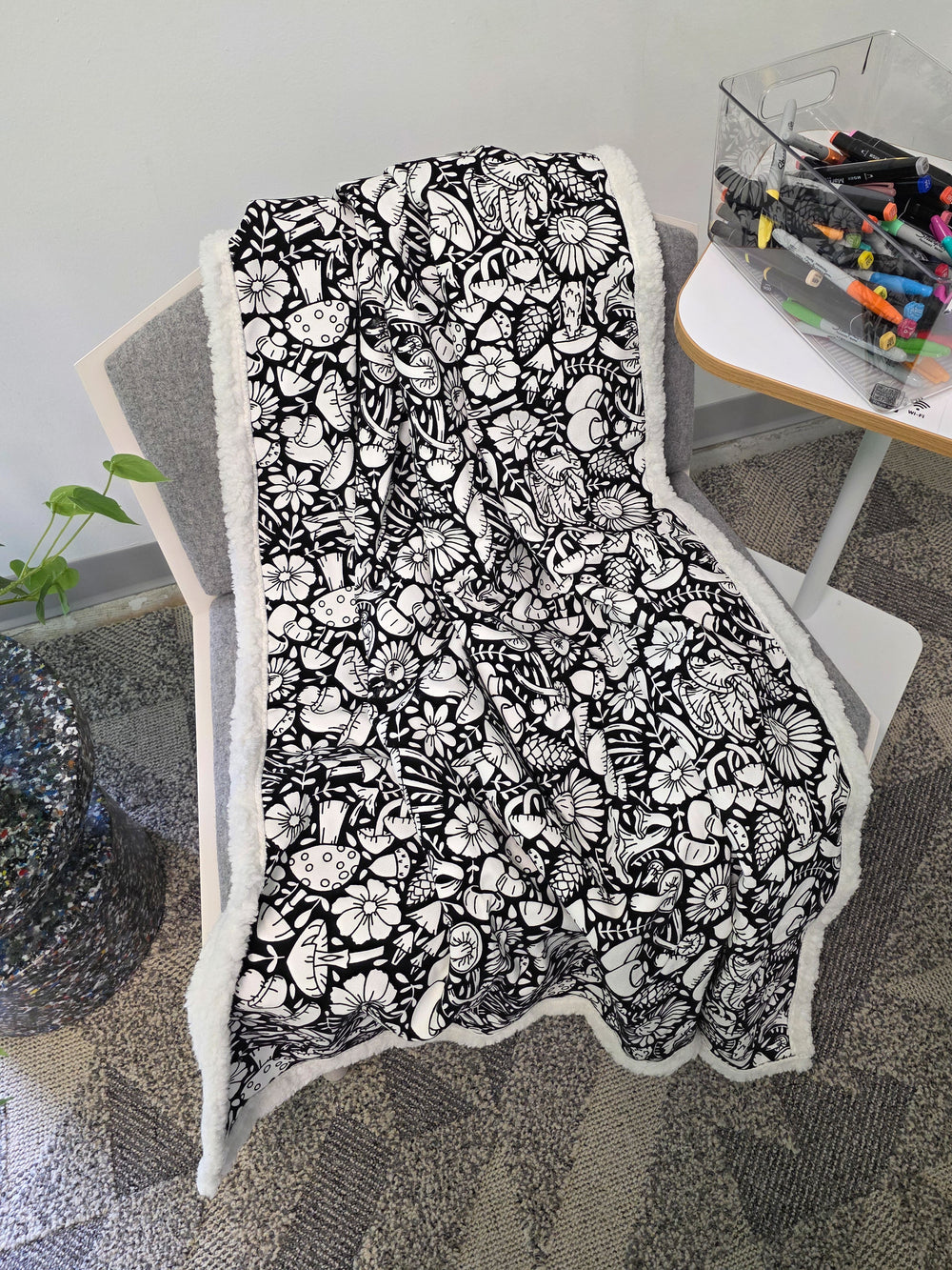 Mushroom color your own blanket draped messily on a gray and white chair next to a table with a box of colorful markers. 
