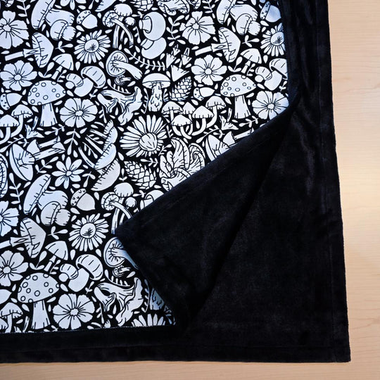 Black and white uncolored mushroom and floral patterned blanket with close up view showing black fleece lined underside