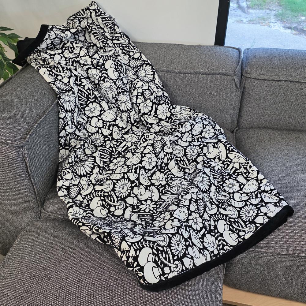 Black and white uncolored mushroom and floral patterned blanket with black fleece lined underside draped over a gray couch.