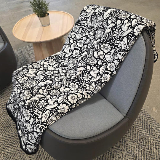Black and white uncolored mushroom and floral patterned blanket with black fleece lined underside draped over a gray chair.