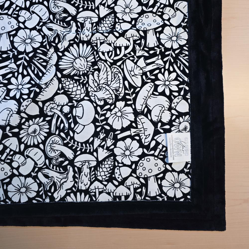 Black and white uncolored mushroom and floral patterned blanket with black fleece lined underside close up view