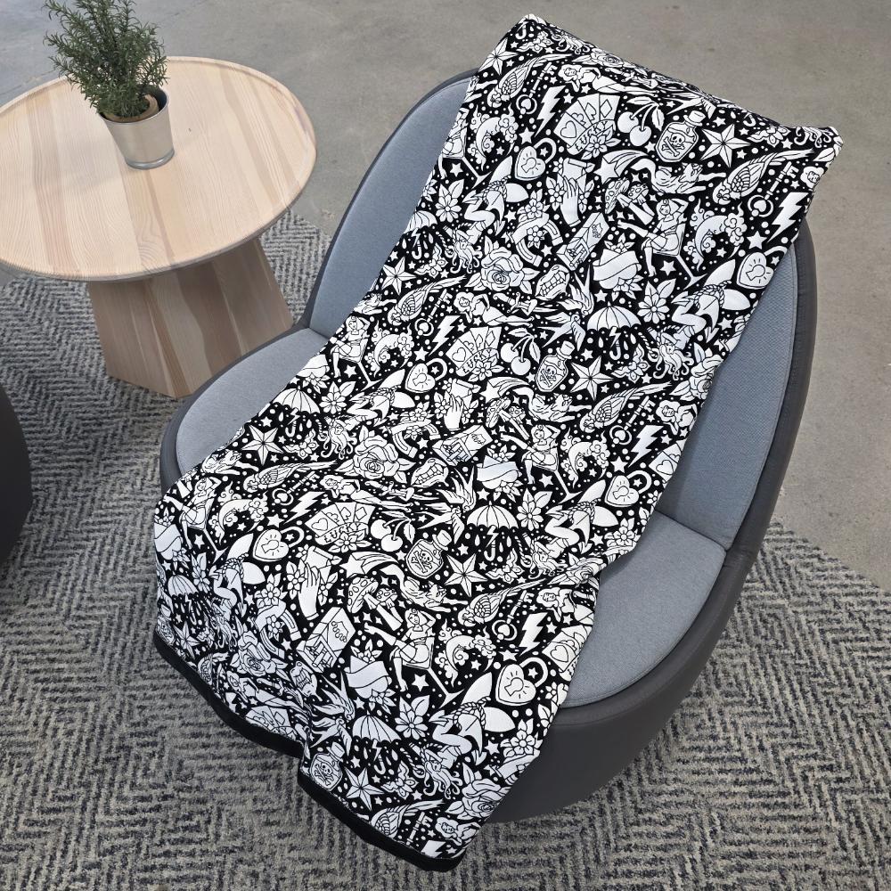 Black and white uncolored classic tattoo styles and patterns blanket with a black fleece lined underside draped over a gray chair.