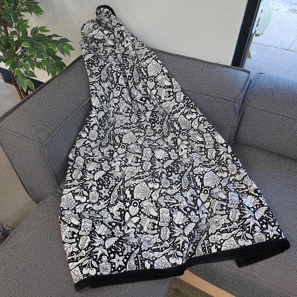Black and white uncolored classic tattoo styles and patterns blanket with a black fleece lined underside draped over a gray couch.