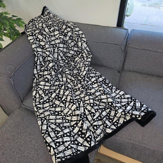 Black and white uncolored many crayon patterned blanket with a black fleece lined underside draped over a gray couch.
