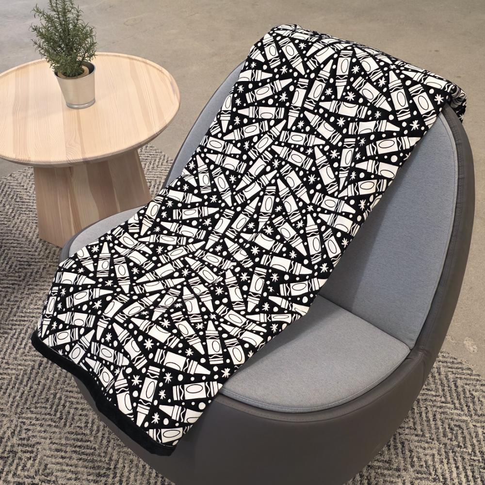 Black and white uncolored many crayon patterned blanket with a black fleece lined underside draped over a gray chair.