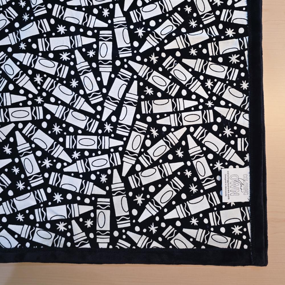 Black and white uncolored many crayon patterned blanket with black fleece lined underside close up view