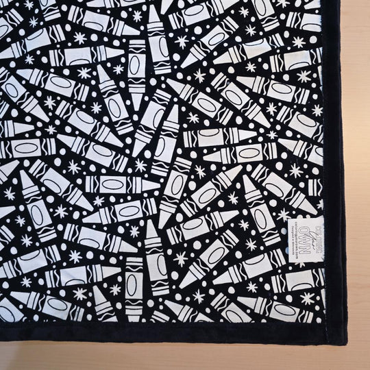 Black and white uncolored many crayon patterned blanket with black fleece lined underside close up view