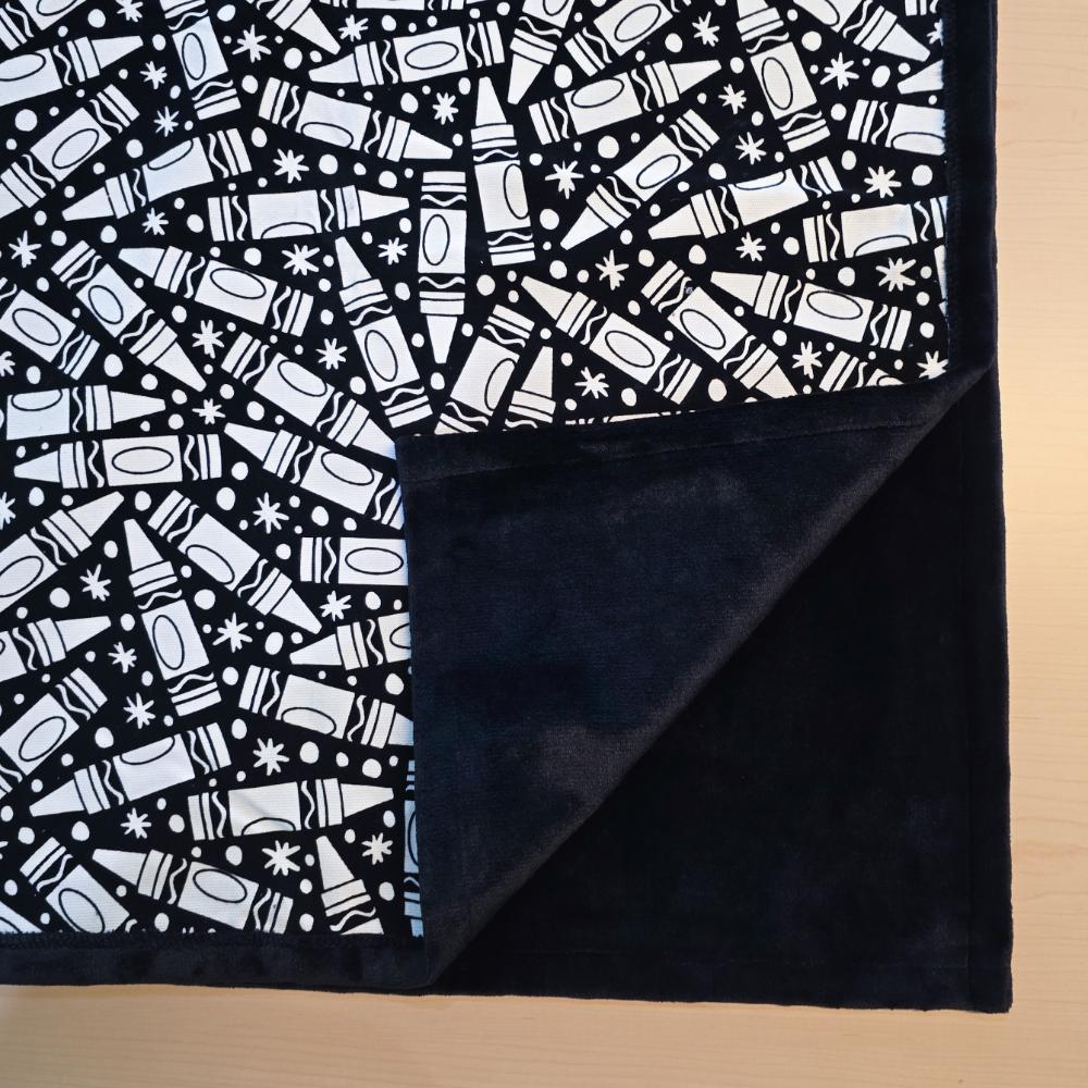 Black and white uncolored many crayon patterned blanket with close up view showing black fleece lined underside