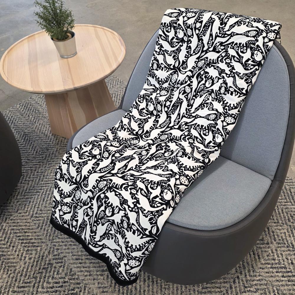 Black and white uncolored dinosaurs with dinosaur eggs patterned blanket with a black fleece lined underside draped over a gray chair.