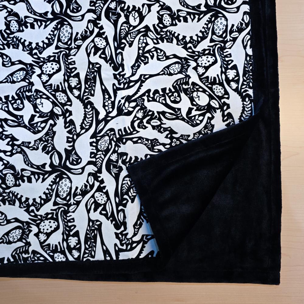 Black and white uncolored dinosaurs with dinosaur eggs patterned blanket with close up view showing black fleece lined underside