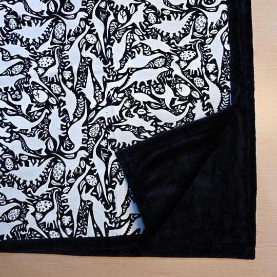 Black and white uncolored dinosaurs with dinosaur eggs patterned blanket with close up view showing black fleece lined underside