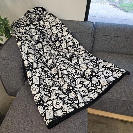 Black and white uncolored version two doodle patterned blanket with donuts, cats, cupcakes a black fleece lined underside draped over a gray couch.