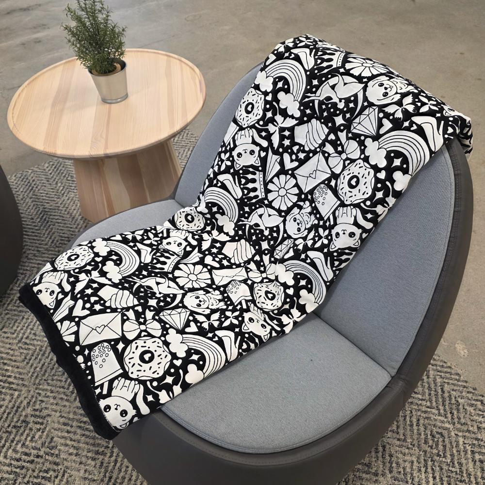 Black and white uncolored version two doodle patterned blanket with donuts, cats, cupcakes  a black fleece lined underside draped over a gray chair.