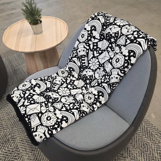 Black and white uncolored version two doodle patterned blanket with donuts, cats, cupcakes  a black fleece lined underside draped over a gray chair.