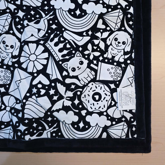 Black and white uncolored version two doodle patterned blanket with donuts, cats, cupcakes  patterned blanket with black fleece lined underside close up view