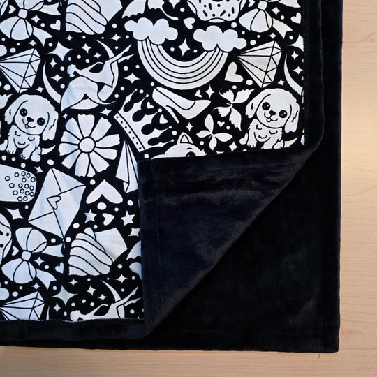 Black and white uncolored version two doodle patterned blanket with donuts, cats, cupcakes with close up view showing black fleece lined underside