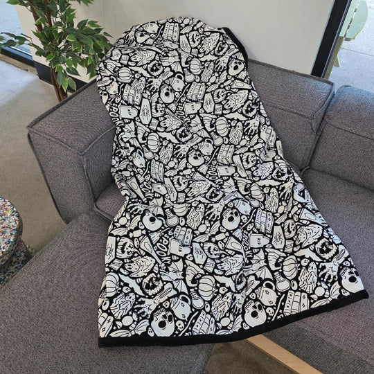 Black and white uncolored spooky halloween patterned blanket with skulls and bats and a black fleece lined underside draped over a gray couch.