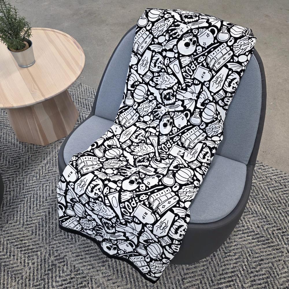 Black and white uncolored spooky halloween patterned blanket with skulls and bats and a black fleece lined underside draped over a gray chair.