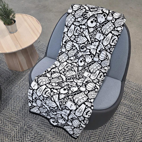 Black and white uncolored spooky halloween patterned blanket with skulls and bats and a black fleece lined underside draped over a gray chair.