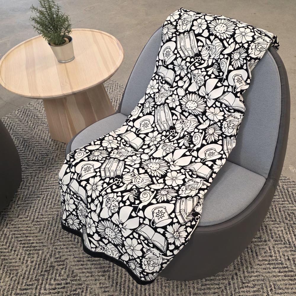 Black and white uncolored highland cow with books and tea patterned blanket with a black fleece lined underside draped over a gray chair.