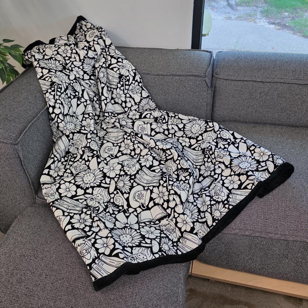 Black and white uncolored highland cow with books and tea patterned blanket with a black fleece lined underside draped over a gray couch.