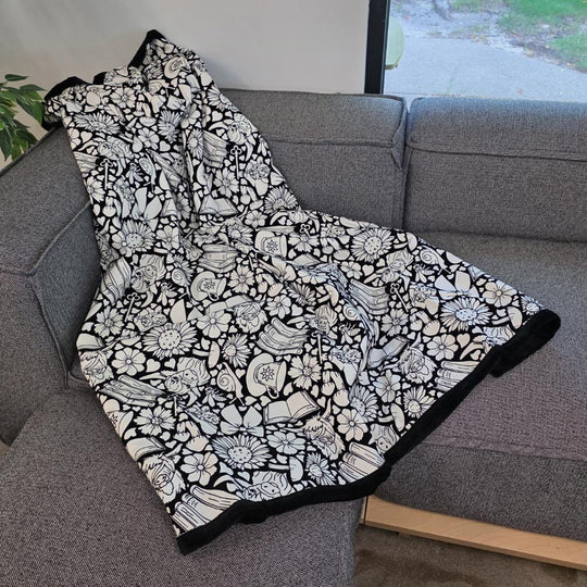 Black and white uncolored highland cow with books and tea patterned blanket with a black fleece lined underside draped over a gray couch.