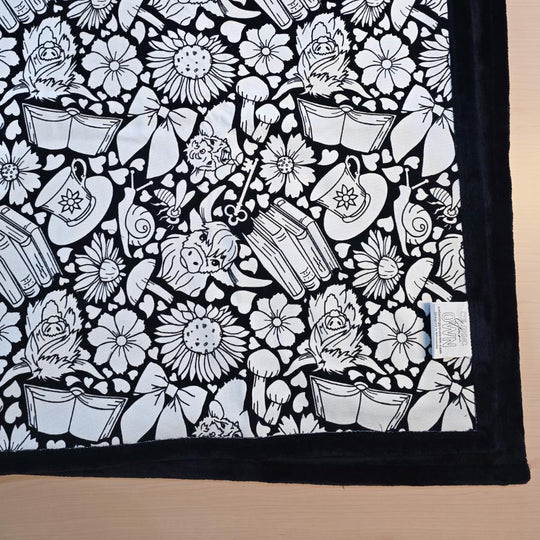 Black and white uncolored highland cow with books and tea  patterned blanket with black fleece lined underside close up view