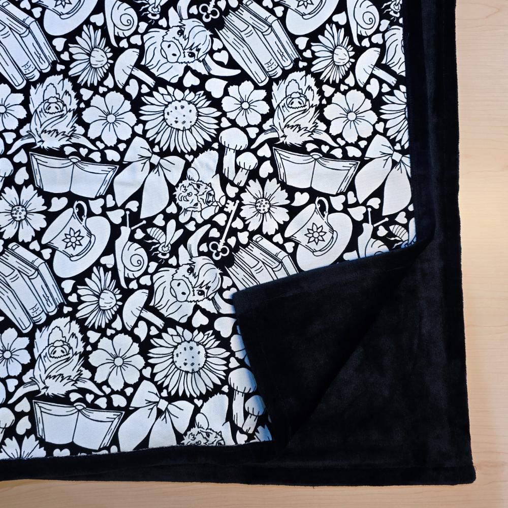 Black and white uncolored highland cow with books and tea patterned blanket with close up view showing black fleece lined underside