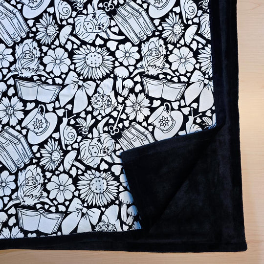 Black and white uncolored highland cow with books and tea patterned blanket with close up view showing black fleece lined underside
