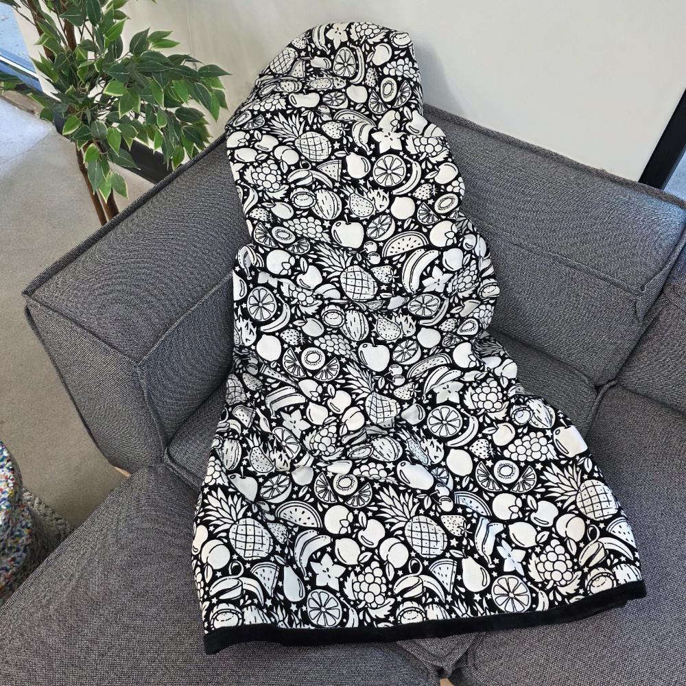 Black and white uncolored fruit variety with pineapples, bananas, watermelons and more patterned blanket with a black fleece lined underside draped over a gray couch.