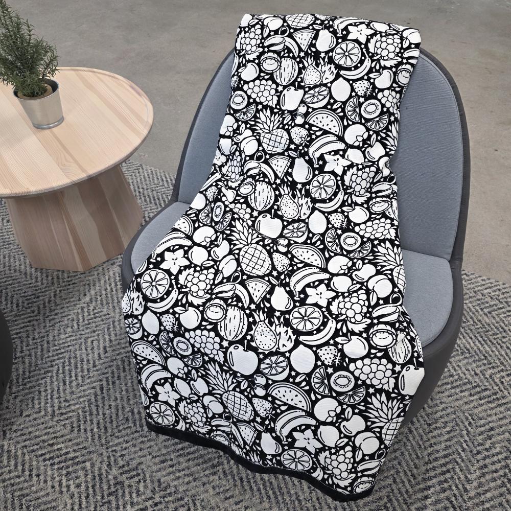 Black and white uncolored fruit variety with pineapples, bananas, watermelons and more patterned blanket with a black fleece lined underside draped over a gray chair.