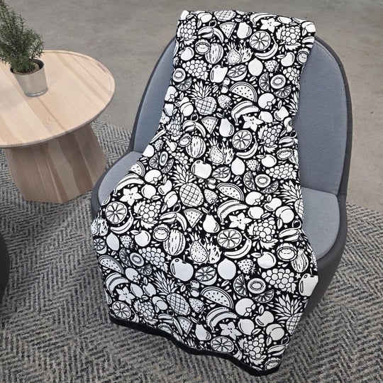 Black and white uncolored fruit variety with pineapples, bananas, watermelons and more patterned blanket with a black fleece lined underside draped over a gray chair.