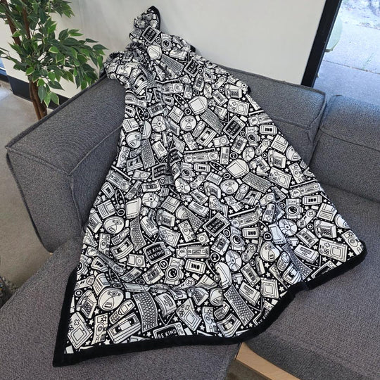 Black and white uncolored retro technology, keyboard ,camera and stereo patterned blanket with a black fleece lined underside draped over a gray couch.
