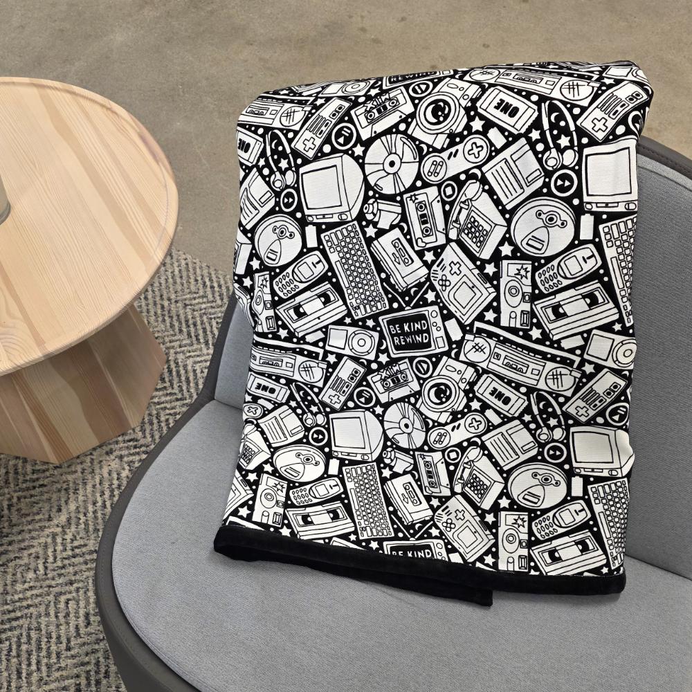 Black and white uncolored retro technology, keyboard ,camera and stereo patterned blanket with a black fleece lined underside draped over a gray chair.