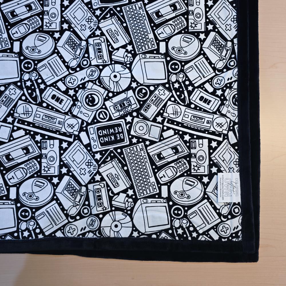 Black and white uncolored retro technology, keyboard ,camera and stereo patterned blanket with black fleece lined underside close up view