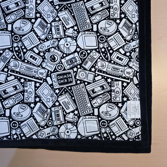 Black and white uncolored retro technology, keyboard ,camera and stereo patterned blanket with black fleece lined underside close up view