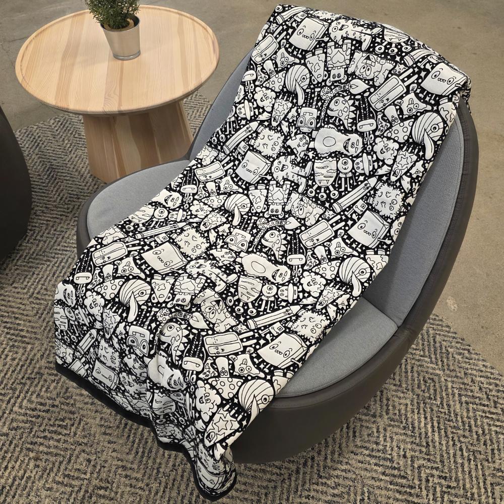 Black and white uncolored silly monster party patterned blanket with black fleece lined underside draped over a gray chair.