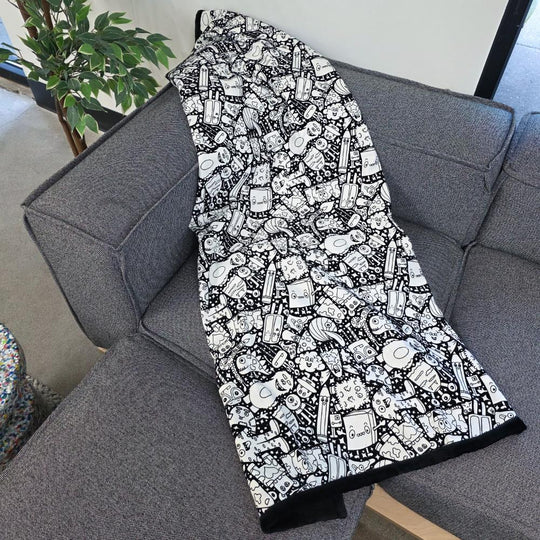 Black and white uncolored silly monster party patterned blanket with black fleece lined underside draped over a gray couch.
