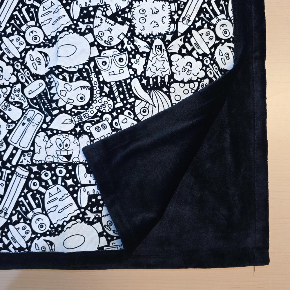 Black and white uncolored silly monster party patterned blanket with close up view showing black fleece lined underside