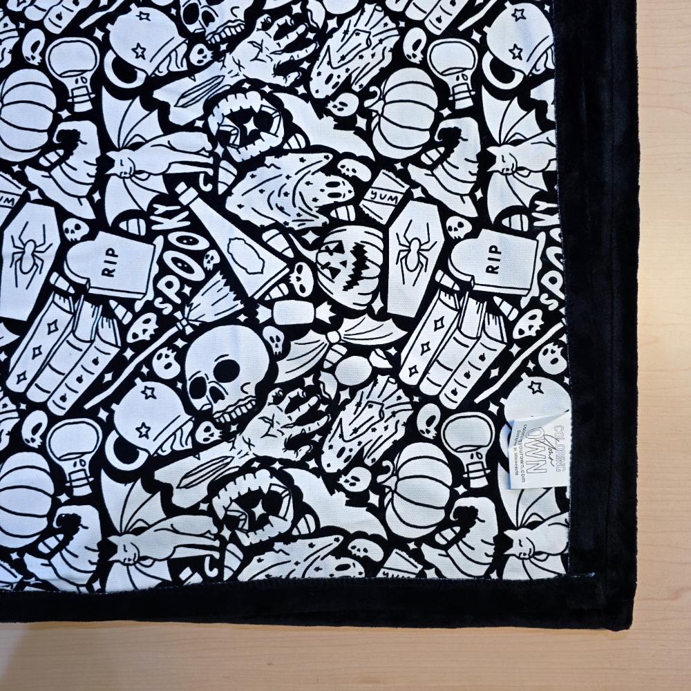 Black and white uncolored spooky Halloween patterned blanket with skulls, ghosts and pumpkins with black fleece lined underside close up view