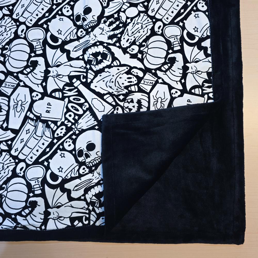Black and white uncolored spooky Halloween patterned blanket with skulls, ghosts and pumpkins with close up view showing black fleece lined underside