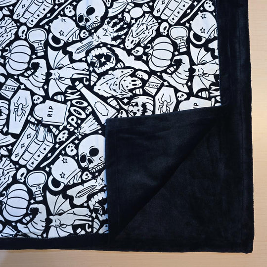 Black and white uncolored spooky Halloween patterned blanket with skulls, ghosts and pumpkins with close up view showing black fleece lined underside