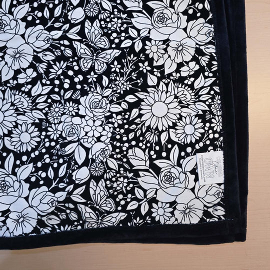 Black and white uncolored vintage floral and butterfly patterned blanket with black fleece lined underside close up view