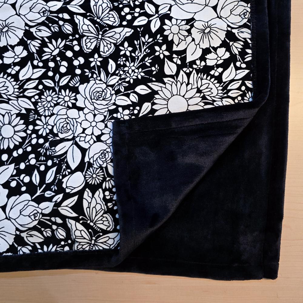 Black and white uncolored vintage floral and butterfly patterned blanket with close up view showing black fleece lined underside