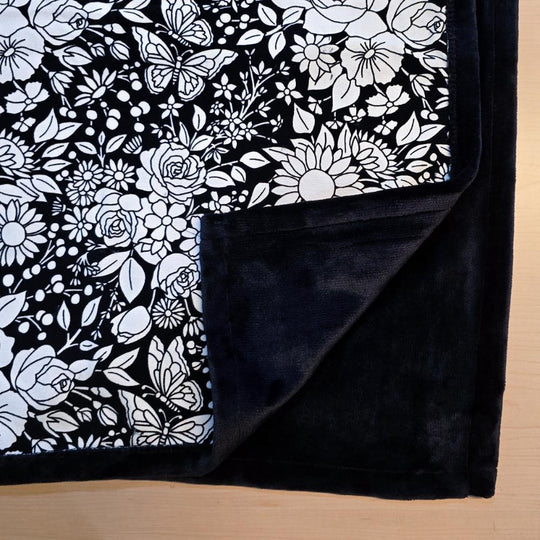 Black and white uncolored vintage floral and butterfly patterned blanket with close up view showing black fleece lined underside